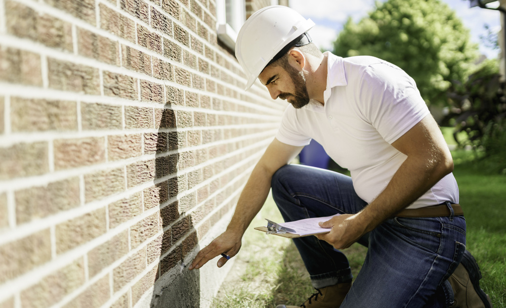 A man with a white hard hat holding a clipboard, inspecting house from the outside walls