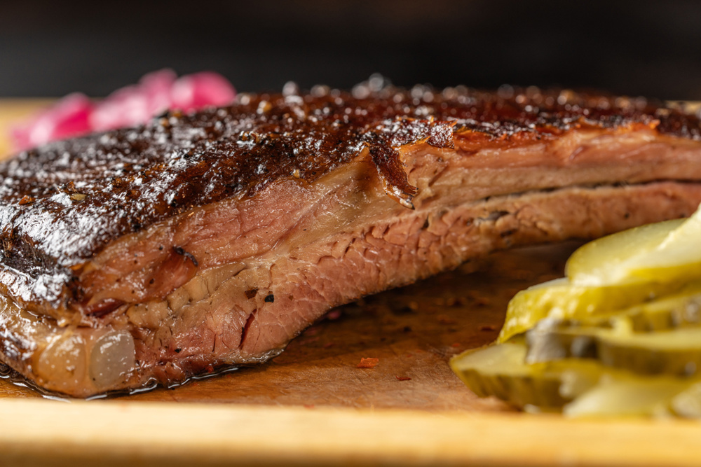 Closeup of tender smoked beef brisket with crispy bark and juicy texture served with pickled cucumbers on wooden board